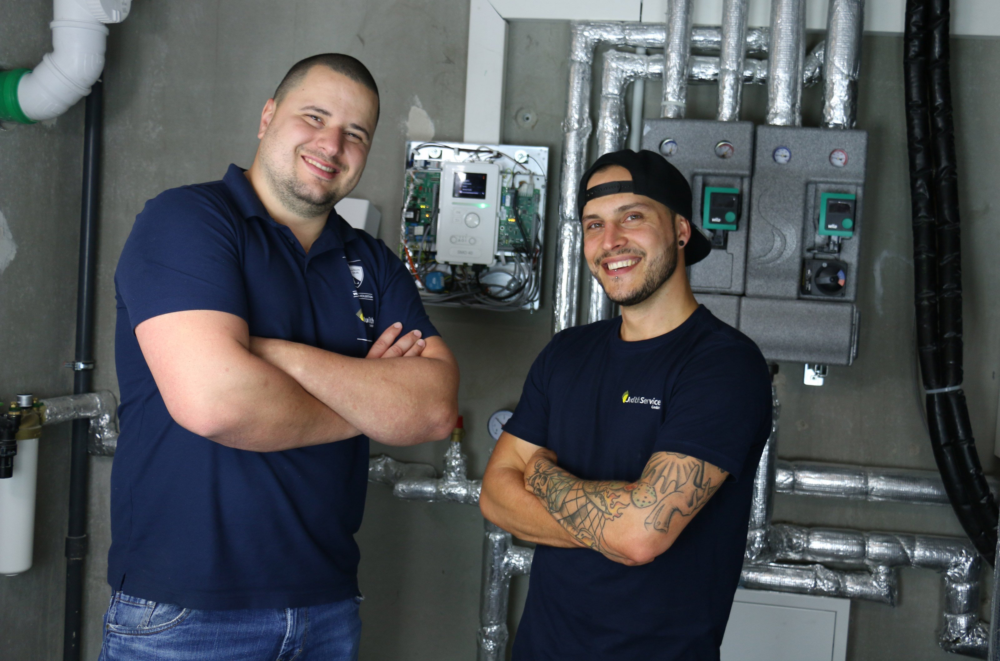 Zwei Männer in blauen T-Shirts stehen lächelnd vor einer technischen Installation mit Rohren und Steuergeräten.