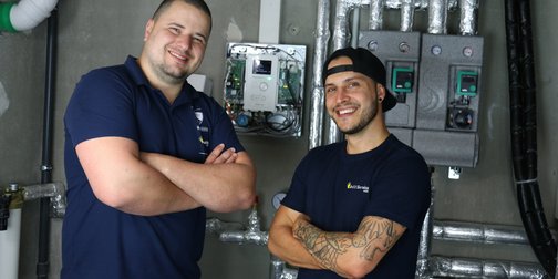 Zwei Techniker lächeln stolz vor moderner Ausrüstung Zwei Männer in blauen T-Shirts stehen lächelnd vor einer technischen Installation mit Rohren und Steuergeräten.