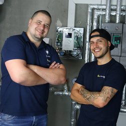 Zwei Techniker lächeln stolz vor moderner Ausrüstung Zwei Männer in blauen T-Shirts stehen lächelnd vor einer technischen Installation mit Rohren und Steuergeräten.