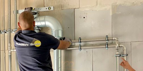 Handwerker bei der Installation von Rohrleitungen in einem Gebäude Ein Handwerker steht auf einer Leiter und installiert Rohre an einer Wand, während seine Kollegen aufmerksam zuschauen.