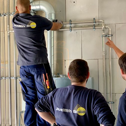 Ein Handwerker steht auf einer Leiter und installiert Rohre an einer Wand, während seine Kollegen aufmerksam zuschauen.