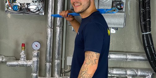Ein junger Mann mit einem weißen Helm und einem blauen Werkzeug in der Hand steht vor einer Wand mit Rohren und Messgeräten.