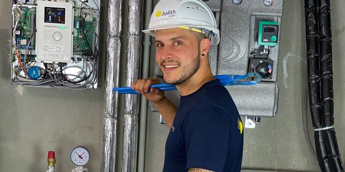 Handwerker mit Helm und Werkzeug in einem modernen Gebäude Ein junger Mann mit einem weißen Helm und einem blauen Werkzeug in der Hand steht vor einer Wand mit Rohren und Messgeräten.