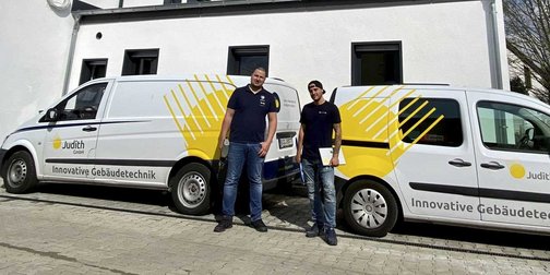 Zwei Techniker vor ihren Fahrzeugen in der Stadt Zwei Techniker stehen vor ihren Fahrzeugen, die mit dem Logo von Judith GmbH und dem Slogan 'Innovative Gebäudetechnik' versehen sind.