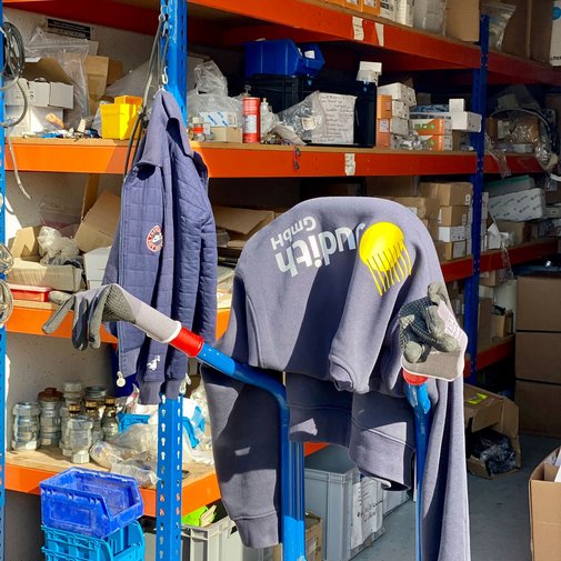 Im Hintergrund sind Regale mit verschiedenen Materialien zu sehen, während ein blauer Wagen mit einem Hoodie und Arbeitshandschuhen gefüllt ist.