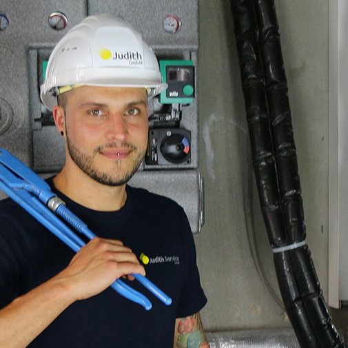 Ein Handwerker mit einem blauen Rohrschneider steht in einem technischen Raum, umgeben von Rohren und Steuergeräten.