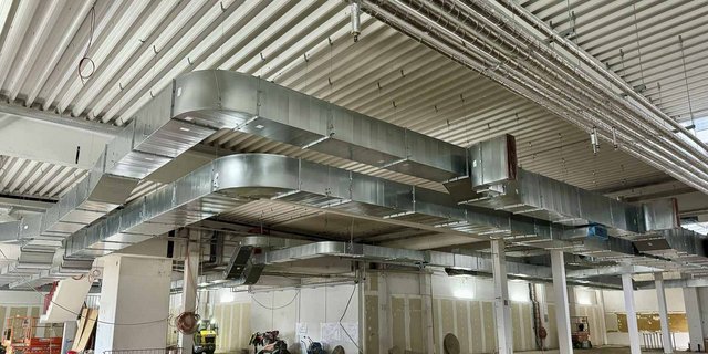 Ein komplexes System von Lüftungskanälen verläuft über die Decke eines großen, unvollendeten Raumes mit weißen Wänden.