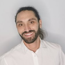 Freundliches Porträt eines Mannes mit Bart und langen Haaren Ein Mann mit langen, lockigen Haaren und einem Bart lächelt freundlich in die Kamera, während er ein weißes Hemd trägt.