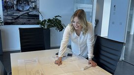 Eine Frau steht an einem Tisch und zeigt auf technische Pläne, während sie in einem modernen Büro lächelt.