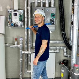 Handwerker in moderner Heizungsanlage mit Werkzeug Ein junger Handwerker mit einem blauen Werkzeug in der Hand lächelt vor einer Wand mit Rohren und technischen Geräten.