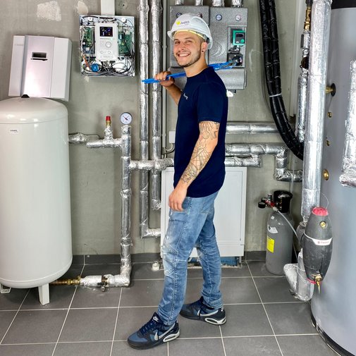 Ein junger Handwerker mit einem blauen Werkzeug in der Hand lächelt vor einer Wand mit Rohren und technischen Geräten.