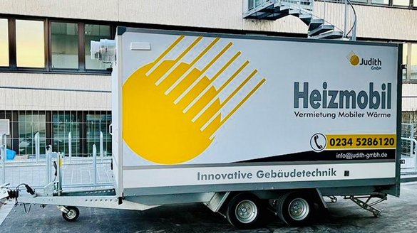 Ein Heizmobil steht vor einem modernen Gebäude, mit einem großen gelben Logo und der Aufschrift 'Vermietung Mobiler Wärme'.