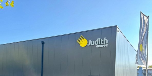 Ein modernes Gebäude mit einer grauen Fassade und dem Logo der Judith Gruppe in gelb und grau, unter einem klaren blauen Himmel.