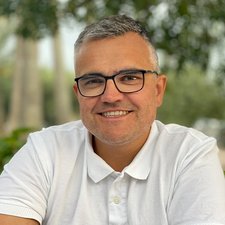 Lächelnder Mann im Freien mit grüner Umgebung Ein lächelnder Mann mit Brille trägt ein weißes Polohemd und sitzt in einer grünen Umgebung mit Bäumen im Hintergrund.