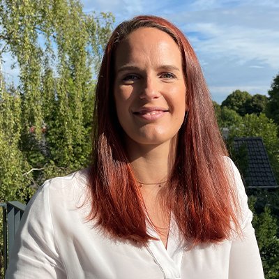 Eine Frau mit langen, roten Haaren lächelt freundlich vor einer grünen, bewaldeten Kulisse und einem blauen Himmel.