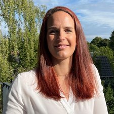 Strahlendes Lächeln einer Frau im Freien Eine Frau mit langen, roten Haaren lächelt freundlich vor einer grünen, bewaldeten Kulisse und einem blauen Himmel.