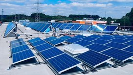 Eine große Fläche mit Solarpanelen auf einem Dach, die in der Sonne glänzen, umgeben von grünen Bäumen und einem blauen Himmel.