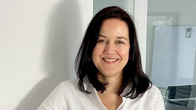 Eine Frau mit langen, dunklen Haaren lächelt freundlich, während sie an einem weißen Schreibtisch steht.