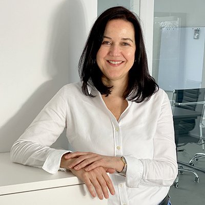Eine Frau mit langen, dunklen Haaren lächelt freundlich, während sie an einem weißen Schreibtisch steht.