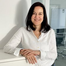 Professionelle Portraitaufnahme einer Frau mit Stil Eine Frau mit langen, dunklen Haaren lächelt freundlich, während sie an einem weißen Schreibtisch steht.