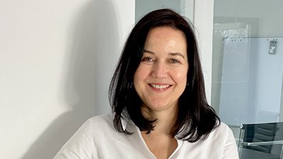 Eine Frau mit langen, dunklen Haaren lächelt freundlich, während sie an einem weißen Schreibtisch steht.