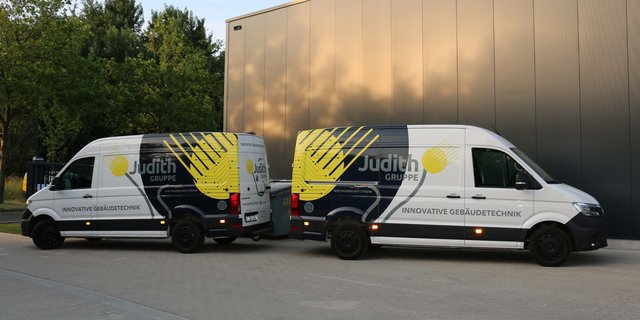 Zwei Lieferwagen der Judith Gruppe stehen auf einem gepflasterten Parkplatz, umgeben von Bäumen und einem modernen Gebäude.
