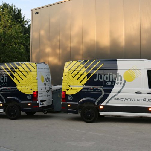 Zwei Lieferwagen der Judith Gruppe stehen auf einem gepflasterten Parkplatz, umgeben von Bäumen und einem modernen Gebäude.