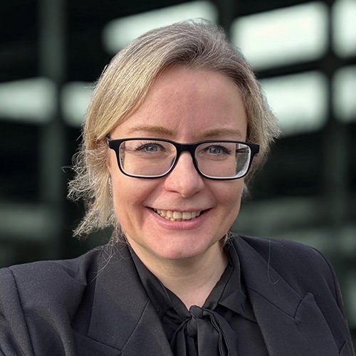 Eine lächelnde Frau mit Brille trägt einen schwarzen Blazer und posiert vor einem modernen Gebäude mit großen Fenstern.