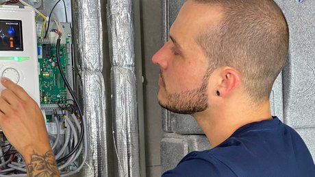 Ein Mann mit einem kurzen Haarschnitt und einem Tattoo an seinem Arm bedient ein technisches Gerät an einer Wand.