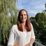 Frau mit rotem Haar genießt den sonnigen Tag Eine Frau mit langen, roten Haaren steht auf einem Balkon und lächelt, umgeben von grünen Bäumen und blauem Himmel.