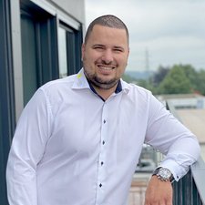 Lächelnder Mann auf Balkon mit urbanem Hintergrund Ein Mann in einem weißen Hemd lächelt entspannt, während er an einem Geländer steht und die Umgebung betrachtet.