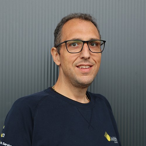Der Mann hat kurzes, lockiges Haar und trägt eine Brille, während er in die Kamera lächelt, umgeben von einer grauen Hintergrundtextur.