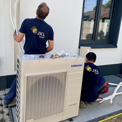 Installation einer Klimaanlage durch Fachleute im Freien Ein Techniker mit einem JKL-GmbH-Shirt richtet Rohre an einer Klimaanlage aus, während ein Kollege im Hintergrund arbeitet.