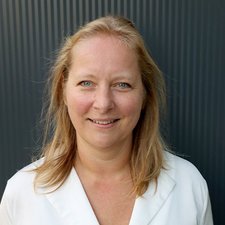 Porträt einer Frau in weißem Oberteil mit natürlichem Licht Eine lächelnde Frau mit blonden Haaren steht vor einer strukturierten, dunklen Wand und strahlt Freundlichkeit aus.