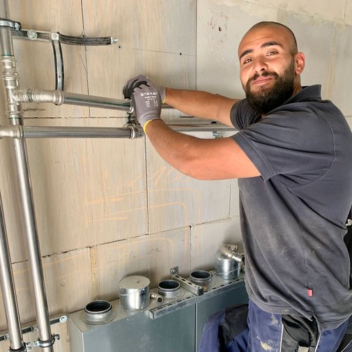 Handwerker installiert Rohre an der Wand mit einem Lächeln Ein Handwerker mit Bart und Handschuhen montiert Rohre an einer Wand, während er lächelt und auf die Kamera schaut.