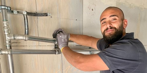 Handwerker installiert Rohre an der Wand mit einem Lächeln Ein Handwerker mit Bart und Handschuhen montiert Rohre an einer Wand, während er lächelt und auf die Kamera schaut.