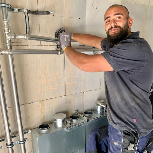 Handwerker installiert Rohre an der Wand mit einem Lächeln Ein Handwerker mit Bart und Handschuhen montiert Rohre an einer Wand, während er lächelt und auf die Kamera schaut.